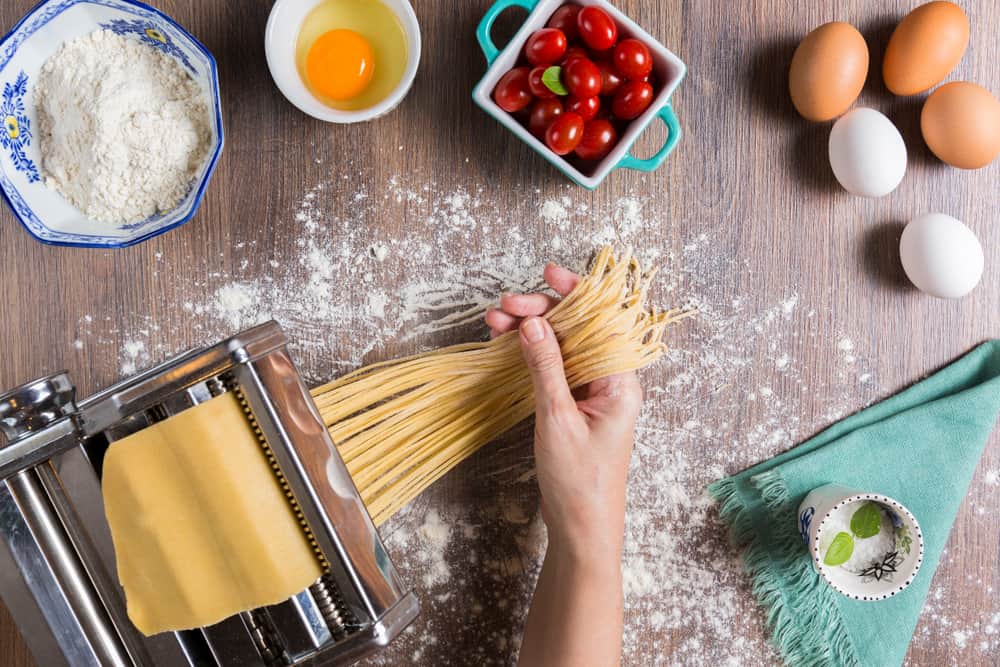 Hænder der trækker strimler af hjemmelavet pasta ud af en pastamaskine. Ved siden af æg, tomater og mel i en skål.