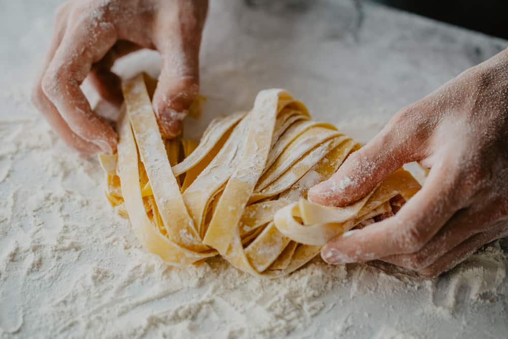 Hænder der holder ved hjemmelavet pasta rullet i mel på marmorplade.
