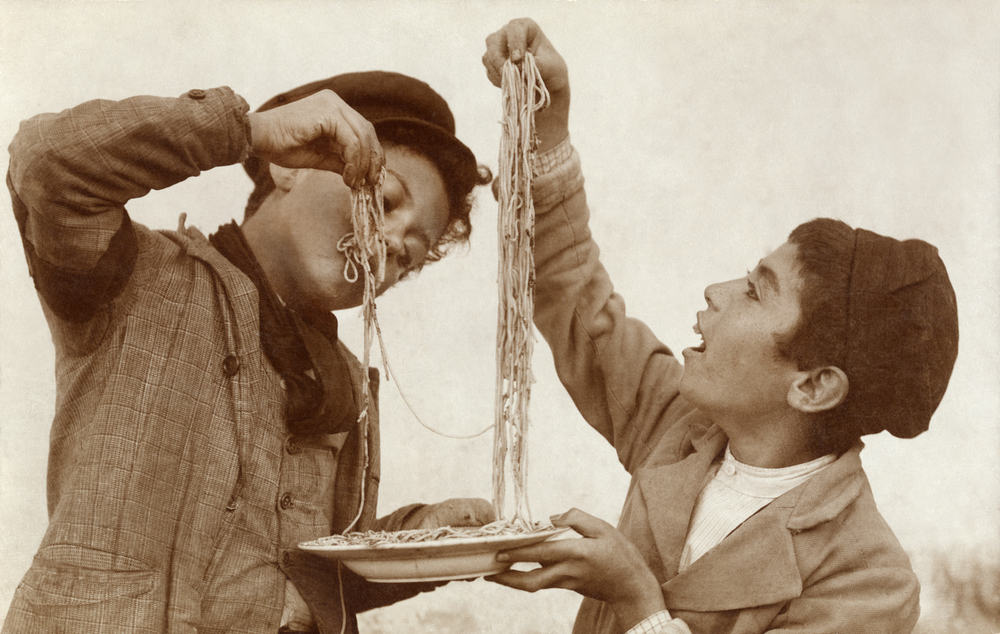 Vintage sort/hvid foto af to drenge der deler en tallerkenfuld spaghetti med fingrene.