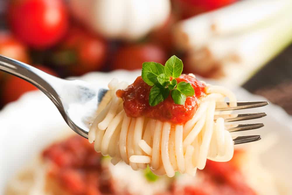 Close up af spisegaffel omviklet af kogt spaghetti. På toppen placeret en smule tomatsovs og lidt grønt.